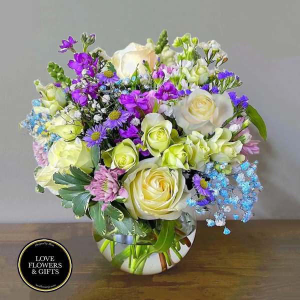 Round glass vase of cream roses, purple blooms, and blue accents in a mixed floral arrangement