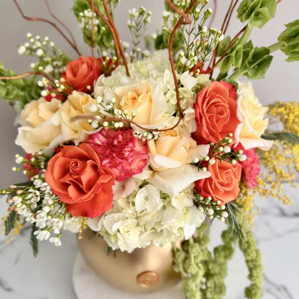 Coral and cream rose arrangement in a gold vase with trailing greenery