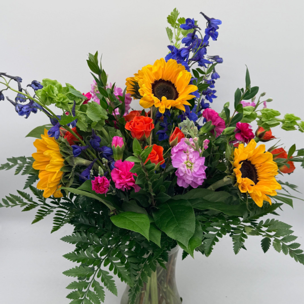 Vibrant mixed bouquet with sunflowers, pink and orange blooms, and blue delphinium in a clear glass vase.