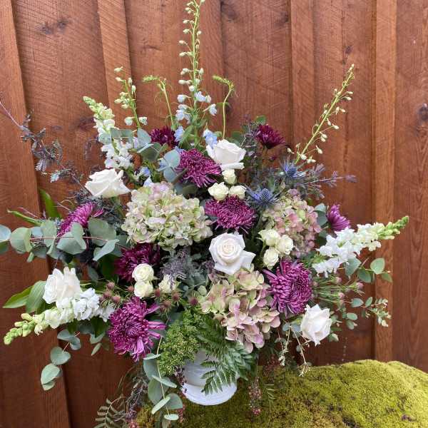 Large floral arrangement with white roses, purple mums, and pale hydrangeas in a white vase.