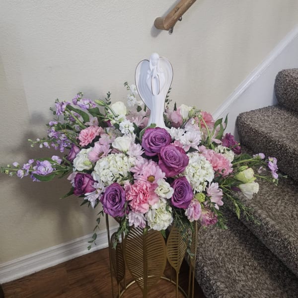 Purple and pink floral arrangement with a white angel figurine on a gold stand
