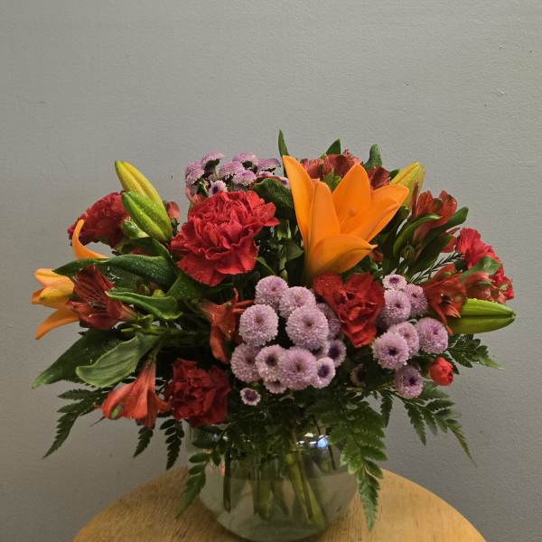 Mixed bouquet of orange lilies, red carnations, and purple mums in a glass vase