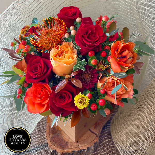 Red and orange rose arrangement with protea and yellow mums in a wooden box on a wood slice base