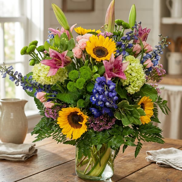 Mixed bouquet with sunflowers, lilies, roses, and blue flowers in a glass vase