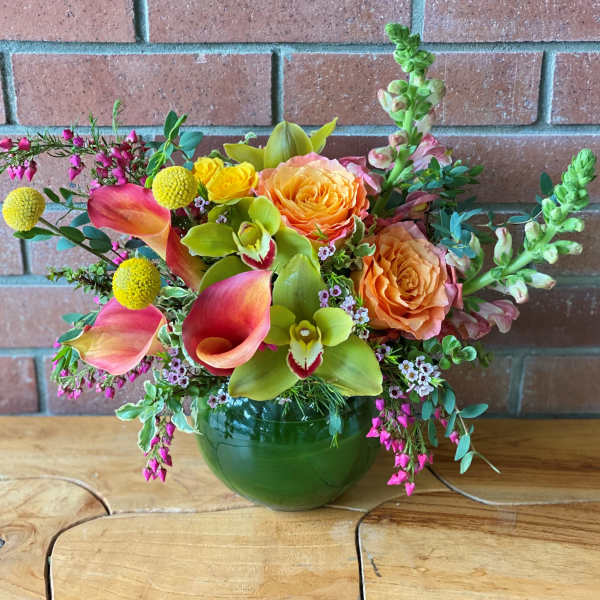 Low arrangement of orange roses, pink calla lilies, and green orchids in a round green glass vase