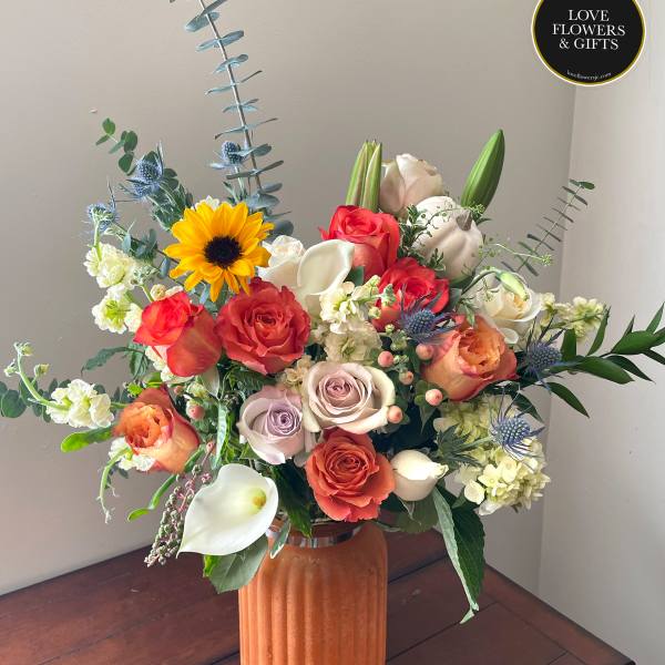 Vibrant mixed arrangement with orange roses, sunflower, white calla lilies and pastel blooms in an orange ribbed vase