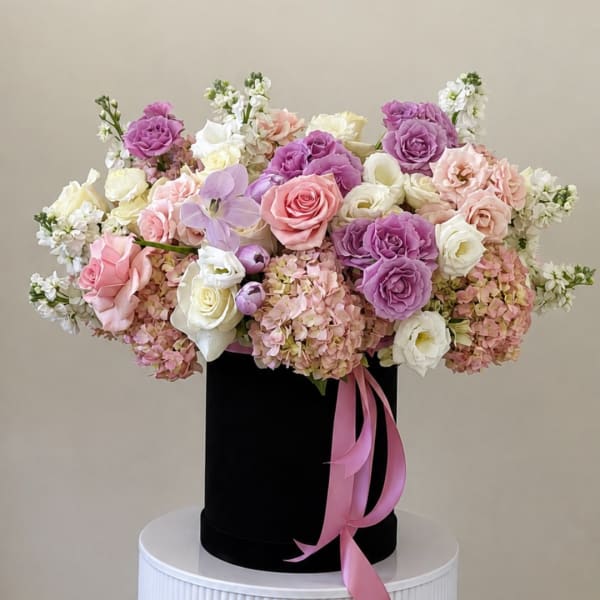 Pink, purple, and white flowers arranged in a black hat box with a pink ribbon.