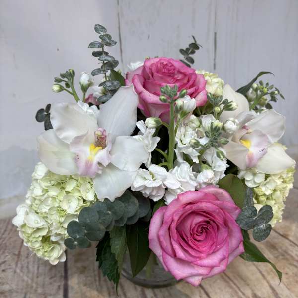 Low arrangement of pink roses, white hydrangeas, stock, and orchids in a clear glass vase