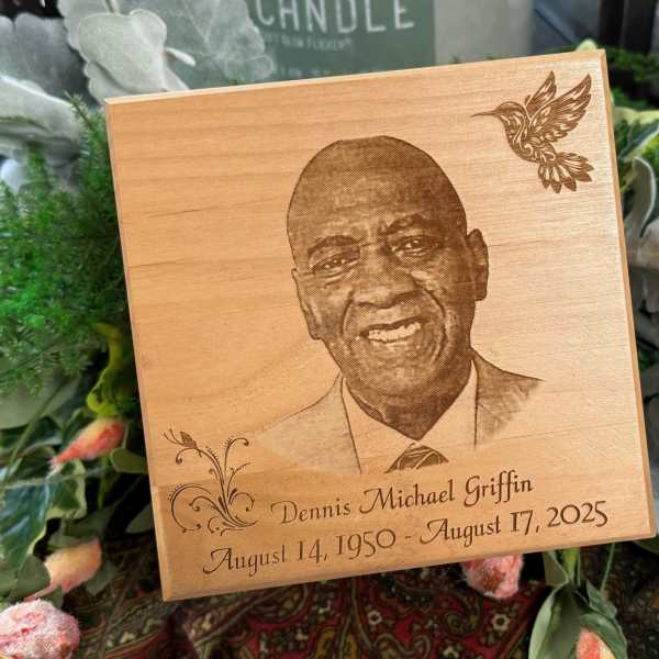Wooden memorial plaque with a portrait and engraved dates, surrounded by flowers