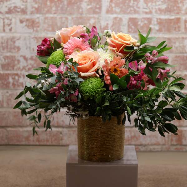 Mixed bouquet of peach roses, pink daisies, and green mums in a gold vase