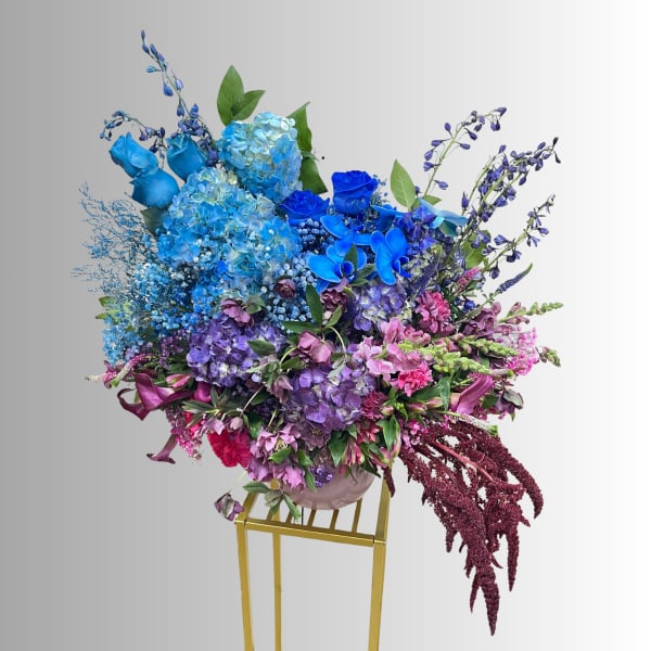 Large blue and purple floral arrangement in a round vase on a gold stand