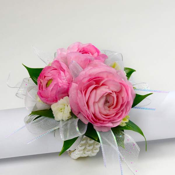Pink ranunculus wrist corsage with white accent flowers and sheer ribbon on a pearl bracelet.
