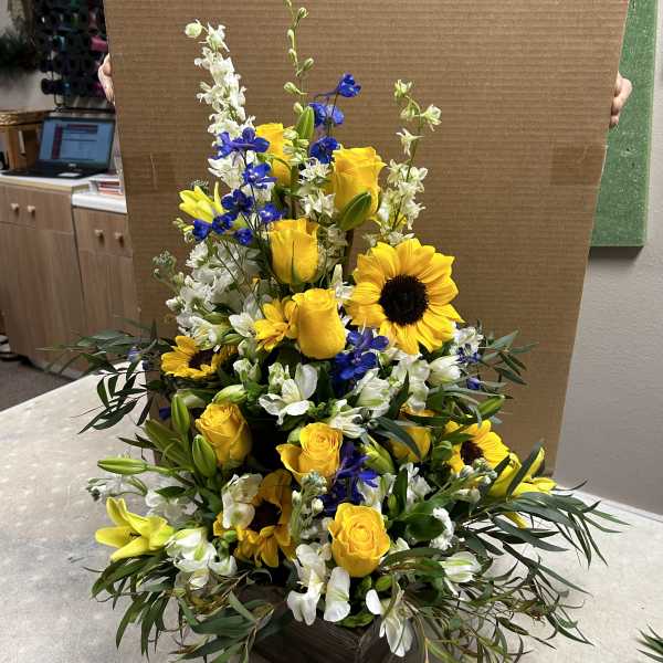Tall floral arrangement with yellow roses, sunflowers, and white blooms in a dark box.