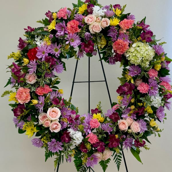 Heart-shaped floral wreath with pink, purple, yellow, and white blooms on a black stand