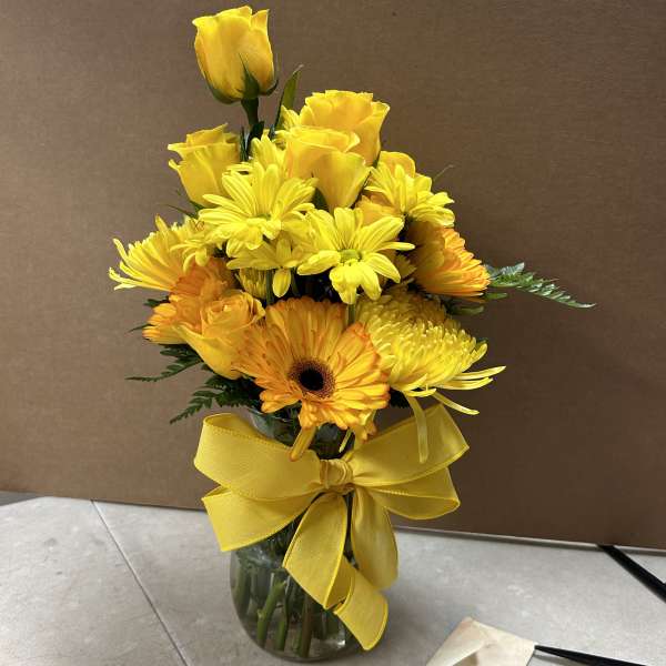 Yellow roses and gerbera daisies in a glass vase with a yellow ribbon