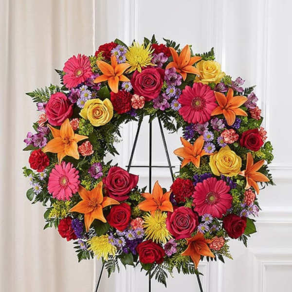 Circular floral wreath on a stand with roses, lilies, gerbera daisies, and small filler flowers