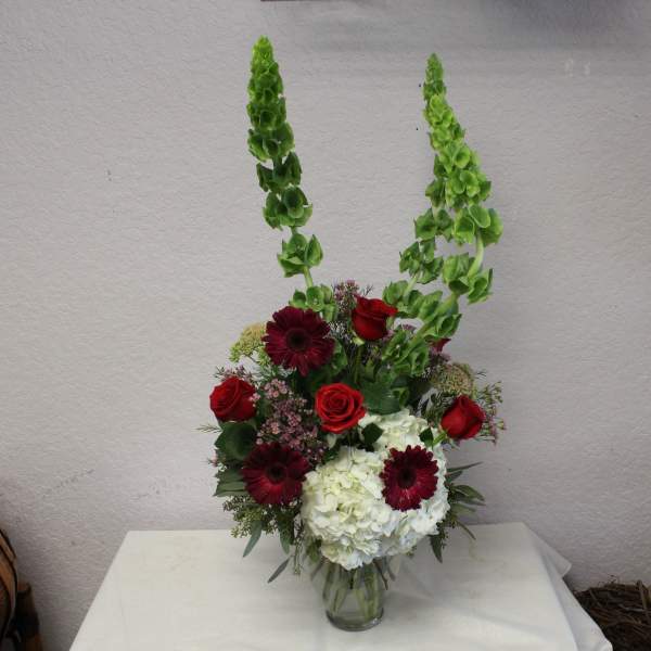 Tall floral arrangement with red roses, burgundy daisies, and white hydrangeas in a glass vase