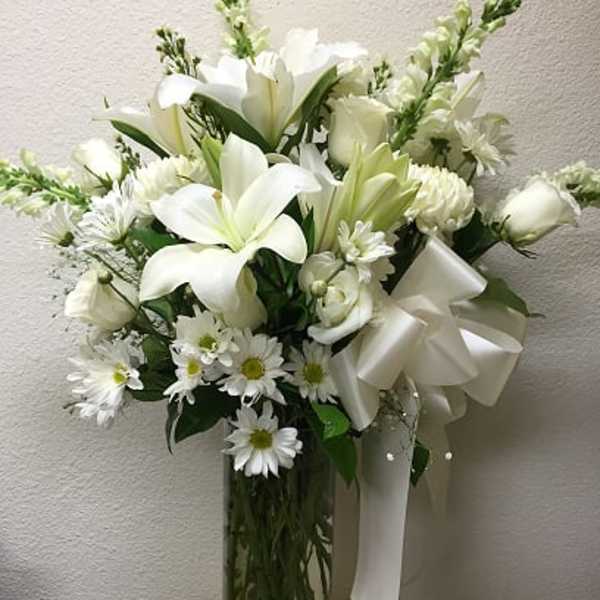 White lilies and daisies arranged in a tall glass vase with a white ribbon.