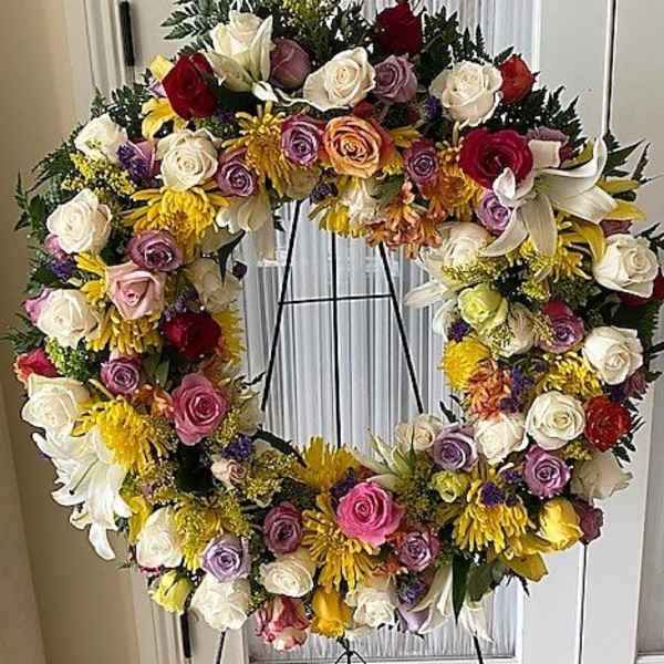 Large circular standing wreath of mixed roses, lilies, and yellow mums on a metal easel stand