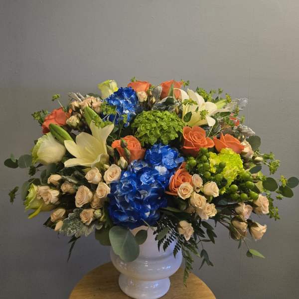 Large mixed bouquet in a white vase with blue, orange, cream, and green blooms