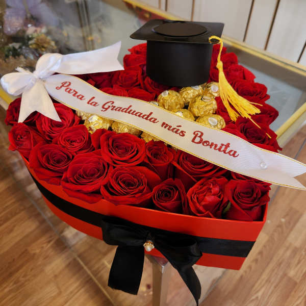 Red roses arranged in a heart-shaped box with gold chocolates and a graduation cap.
