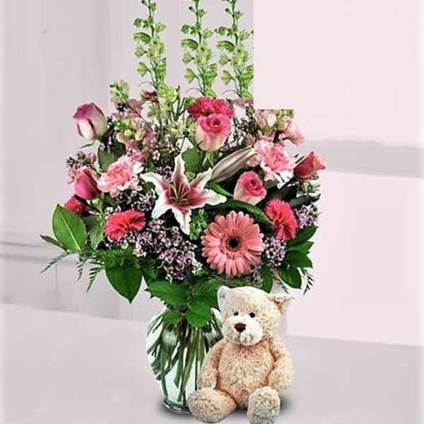 Pink mixed flower arrangement with lilies, roses, and gerbera daisies in a glass vase with a tan teddy bear.