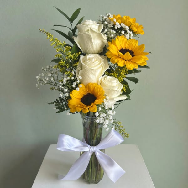 White roses and yellow sunflowers in a clear glass vase with a white ribbon