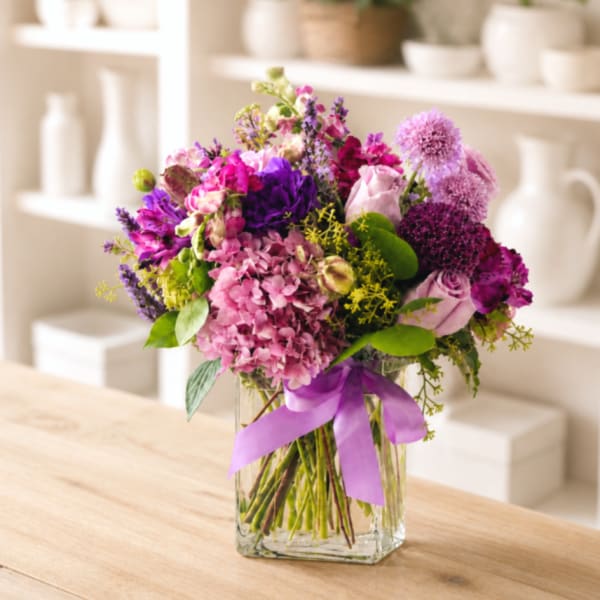 Mixed pink and purple bouquet in a clear square vase with a lavender ribbon