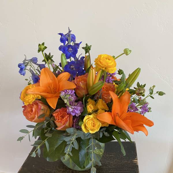 Bright mixed bouquet with orange lilies, roses, and blue flowers in a glass vase
