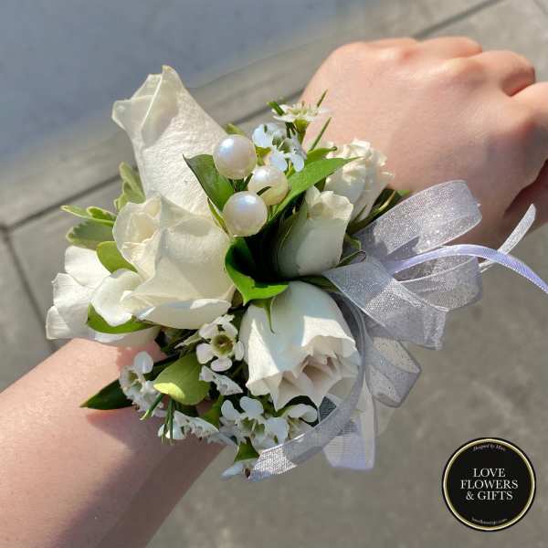 White rose wrist corsage with small white blossoms, pearl accents, and silver ribbon on a person's wrist.