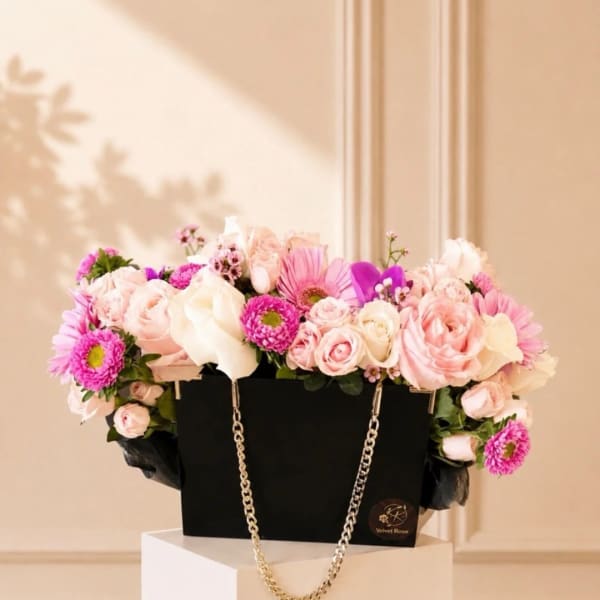 Pink and white flowers arranged in a black handbag-style box with a gold chain