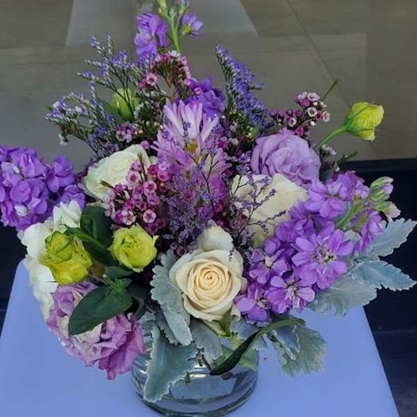 Low glass vase of cream roses and purple mixed flowers on a light blue cube.