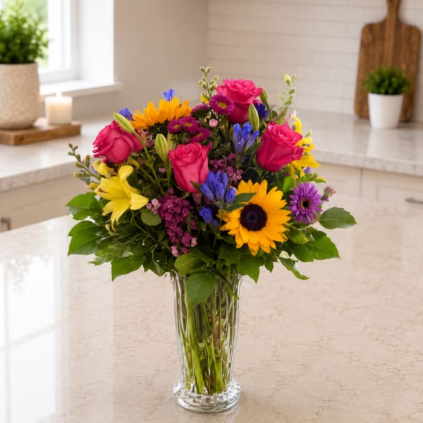 Mixed bouquet of roses, sunflowers, lilies, and purple blooms in a clear glass vase