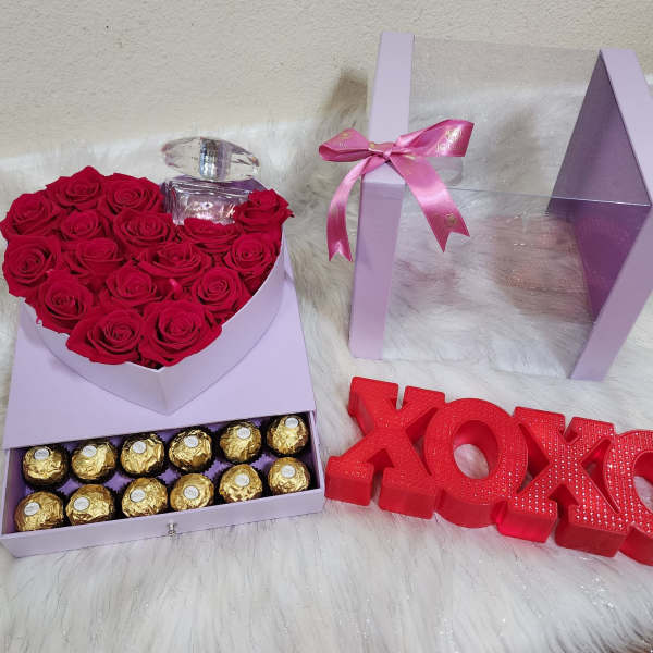 Heart-shaped box of red roses with a perfume bottle and chocolates beside XOXO decor
