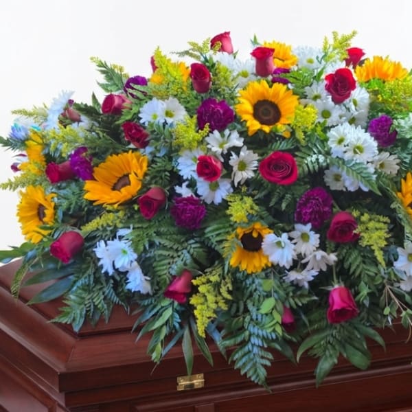 Large floral spray with sunflowers, roses, and white daisies on a wooden casket