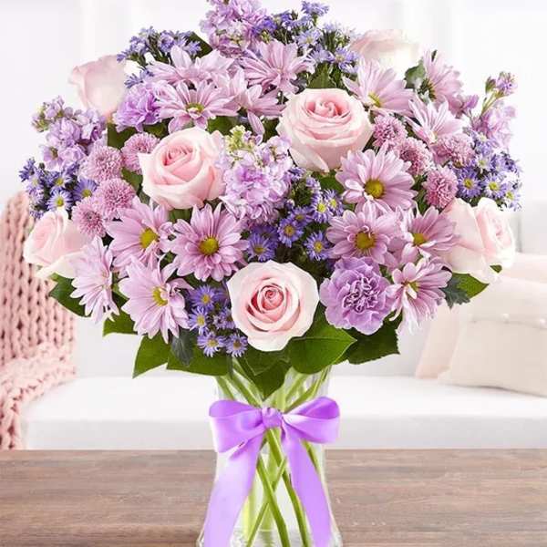 Pink and lavender bouquet of roses and daisies in a glass vase with a purple ribbon