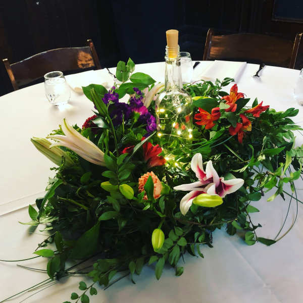 Low floral centerpiece with lilies, orange blooms, and a bottle wrapped in lights