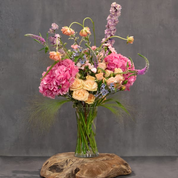 Tall bouquet of pink hydrangeas, peach roses, and lavender flowers in a glass vase
