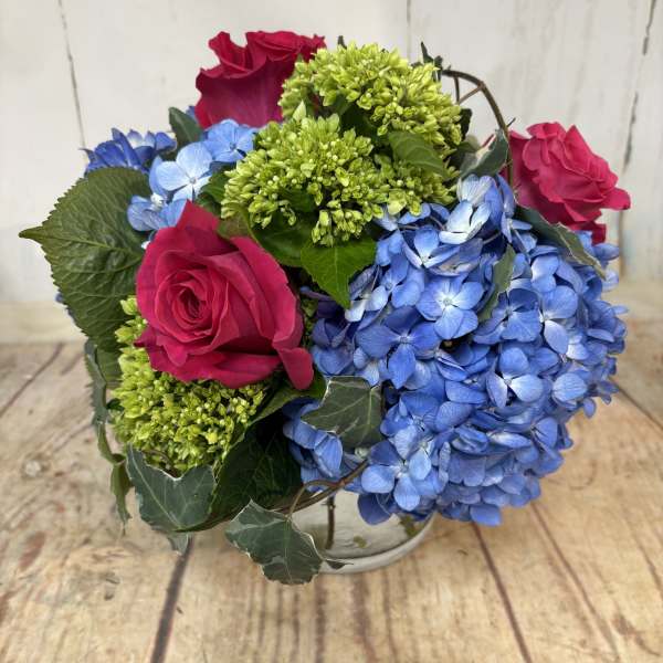 Low round arrangement of hot pink roses, green blooms, and blue hydrangeas in a clear glass vase