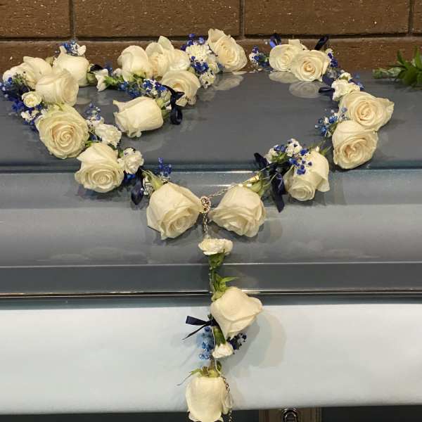 White rose heart-shaped casket spray with blue accents and a hanging rosary