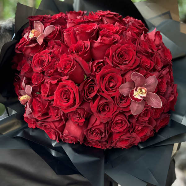 Round bouquet of red roses with a few pink orchids wrapped in black paper