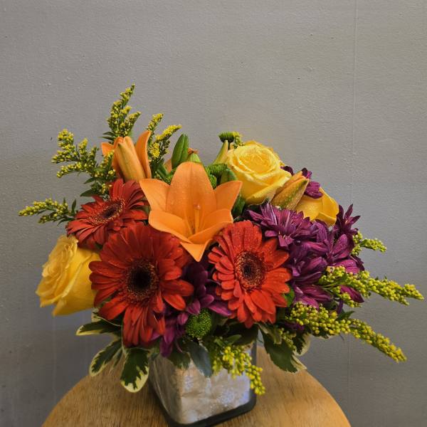 Colorful bouquet with orange lilies, red gerbera daisies, yellow roses, and purple flowers in a square vase