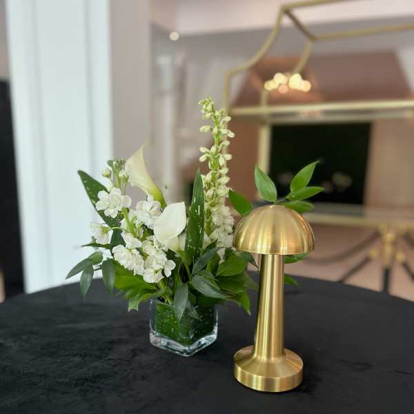 White floral arrangement in a clear square vase beside a gold table lamp