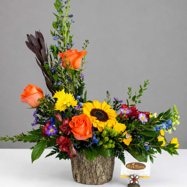 Mixed floral arrangement with orange roses, sunflowers, and blue accents in a textured container