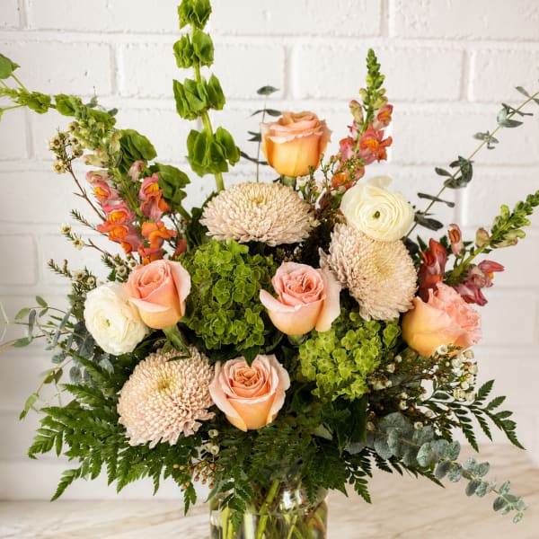 Mixed bouquet of peach roses, cream blooms, and green flowers in a glass vase