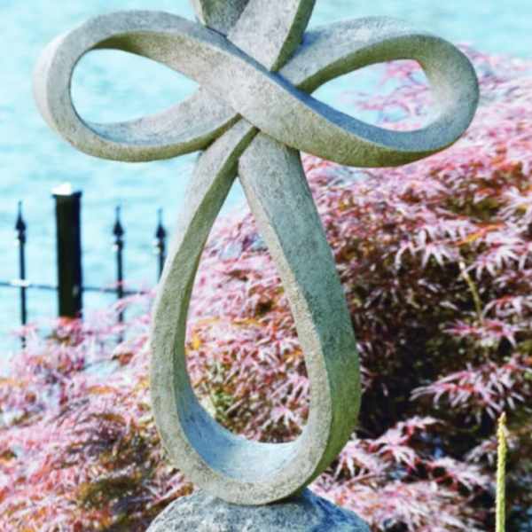Stone sculpture with interlocking loops beside a pond and pink shrubs