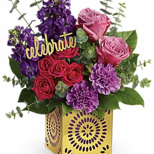 Bright arrangement of pink and red roses with purple flowers in a gold cube vase with a celebrate sign.