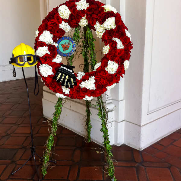 Red and white floral wreath on an easel with a firefighter helmet and gloves
