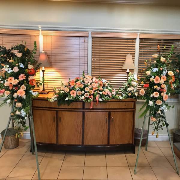 Three floral funeral sprays with peach and white flowers on stands and a casket spray on a cabinet