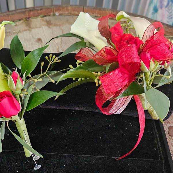 Two calla lily corsages with red ribbon and pink rosebuds on a black box.